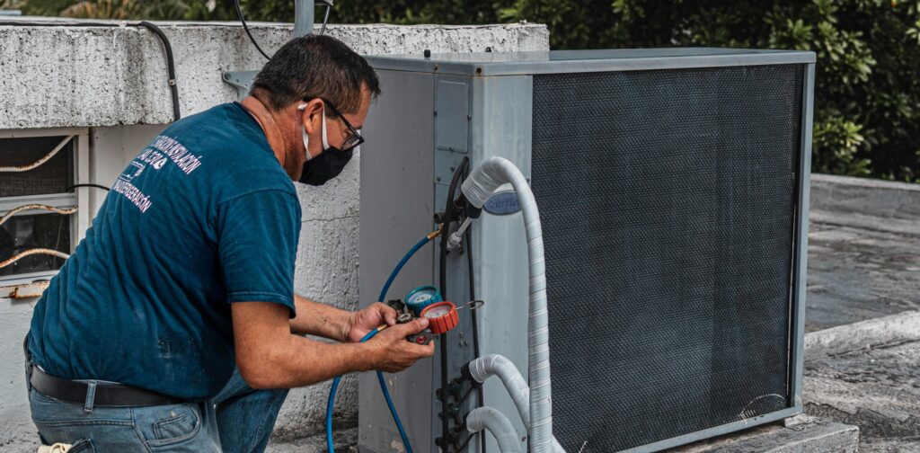 Technician repairing an air conditioner unit outdoors, wearing a facemask and using a manifold gauge.